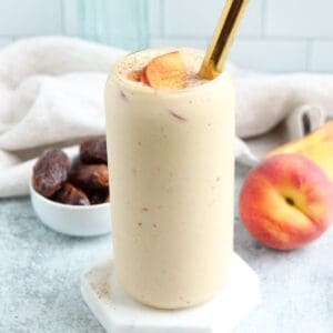 A creamy peach yogurt smoothie in a tall glass, topped with peach slices and a sprinkle of cinnamon. A gold straw rests in the glass. Nearby are a fresh peach and a bowl of dates, with a light cloth in the background.