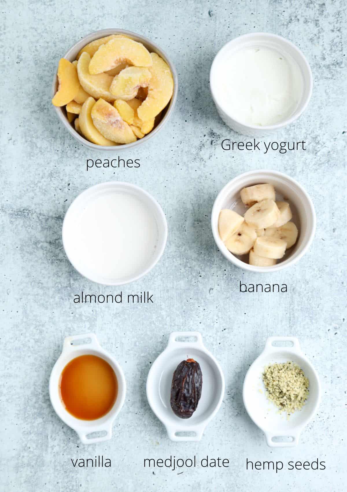 Top-down view of bowls containing ingredients for a peach yogurt smoothie—peaches, Greek yogurt, almond milk, sliced banana, vanilla extract, a medjool date, and hemp seeds—all labeled on a light gray background.