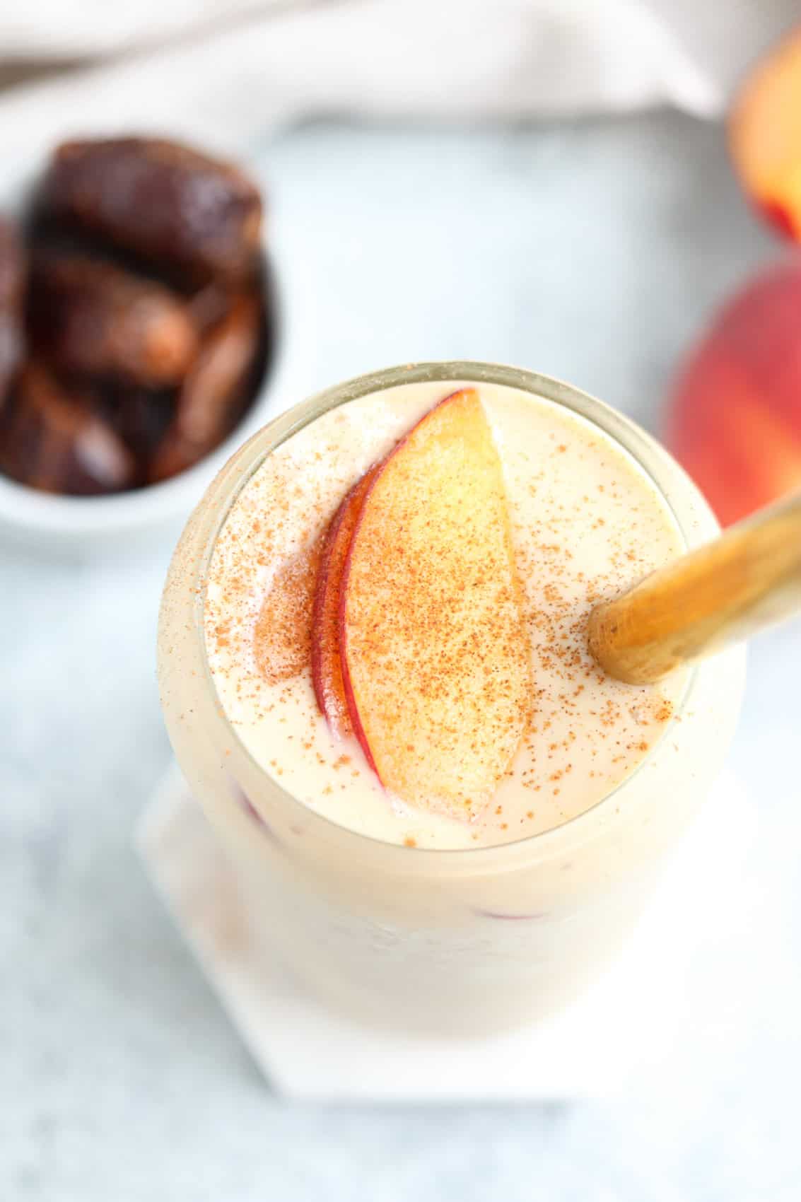 A creamy peach yogurt smoothie in a glass, garnished with peach slices and a sprinkle of cinnamon, with a bamboo straw. In the background, a small bowl of dates and a whole peach are slightly out of focus.