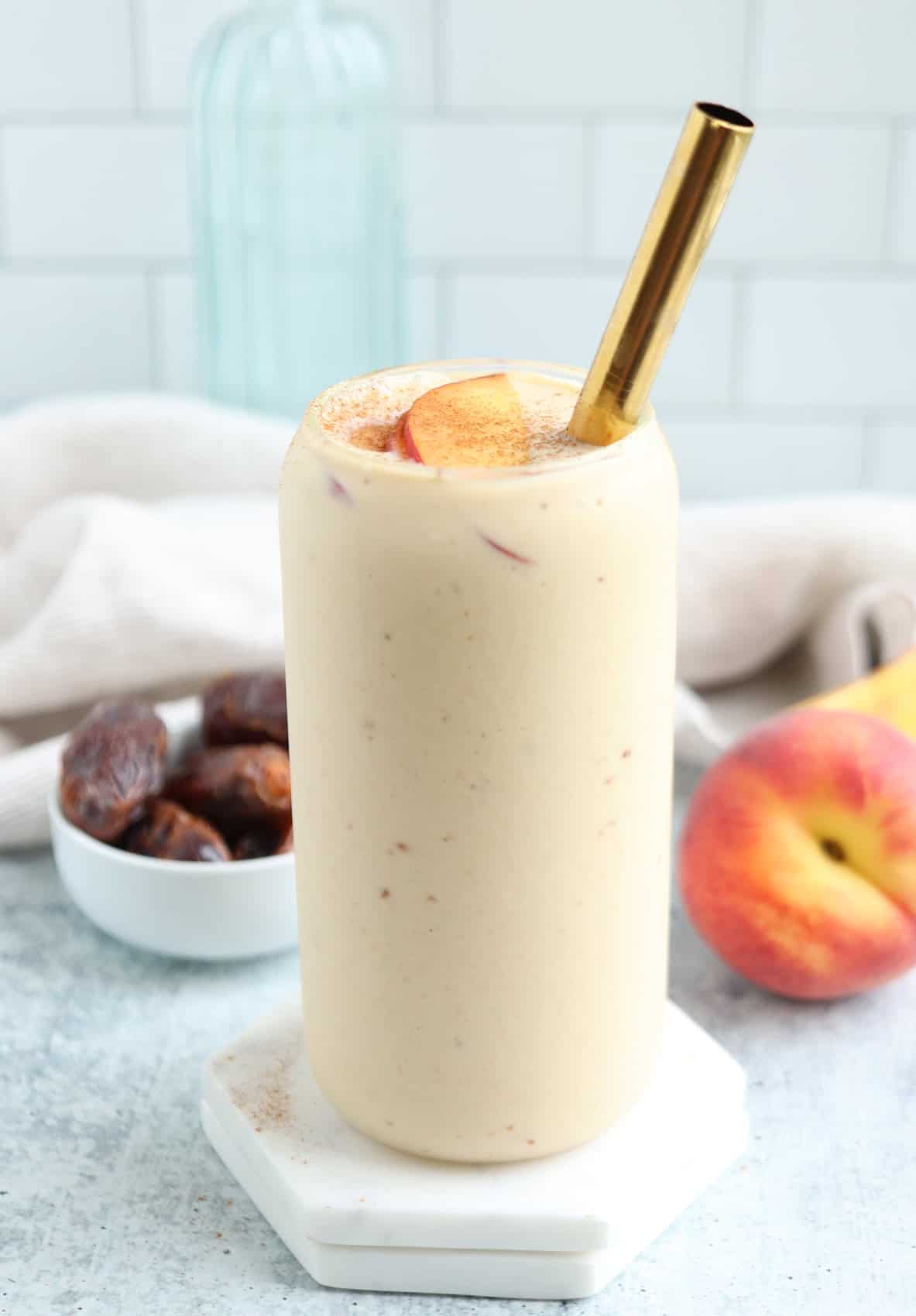 A creamy peach yogurt smoothie in a tall glass with a gold straw, garnished with peach slices and cinnamon. A bowl of dates, a peach, and a light cloth are in the background on a light-colored surface.