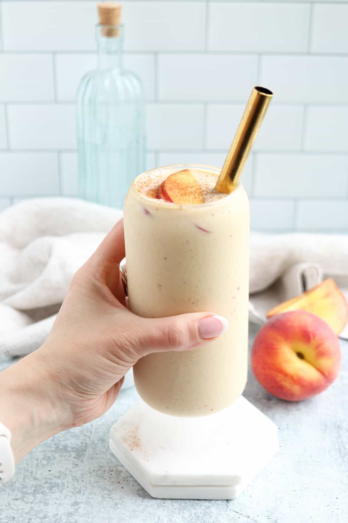 A hand holds a creamy peach yogurt smoothie in a glass with a gold straw, topped with peach slices. A peach and a bottle are in the background, set on a light surface with a tiled wall.