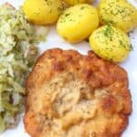 A plate with a breaded pork chop, boiled potatoes topped with dill, and cooked cabbage. Text above reads “Authentic Polish Kotlet Schabowy Breaded Pork Chop,” highlighting the traditional flavors of kotlet schabowy alongside classic sides.