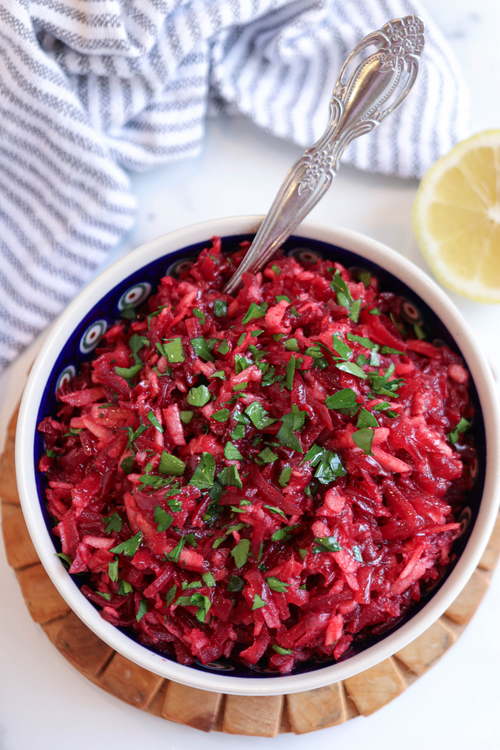 A bowl filled with grated beet and carrot salad, inspired by Polish beet salad, garnished with chopped parsley. A silver spoon rests in the bowl, which is placed on a wooden mat, with a striped cloth and a lemon half nearby.