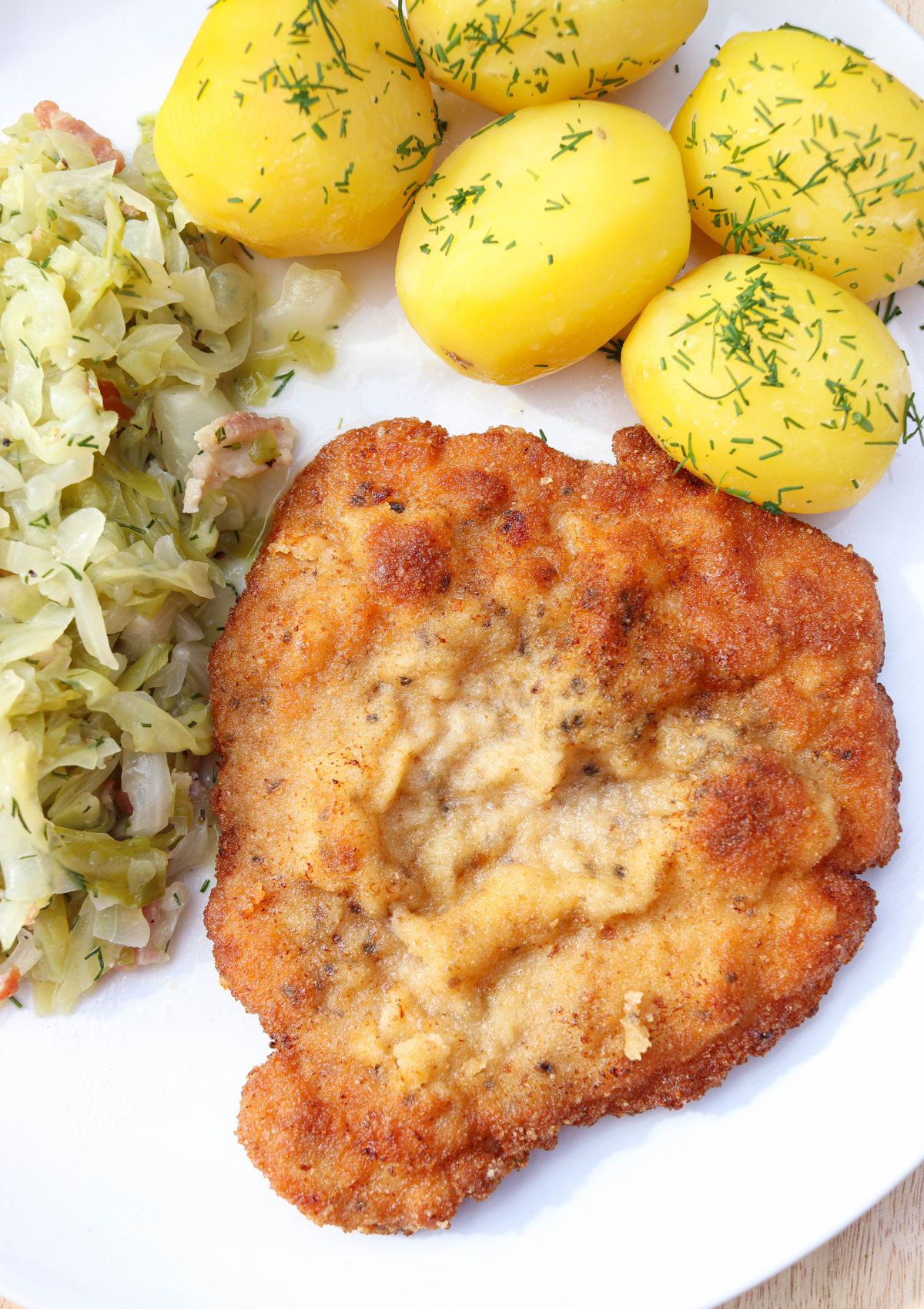 Breaded and fried kotlet schabowy served with boiled potatoes topped with fresh dill and a side of sautéed cabbage with bacon on a white plate.