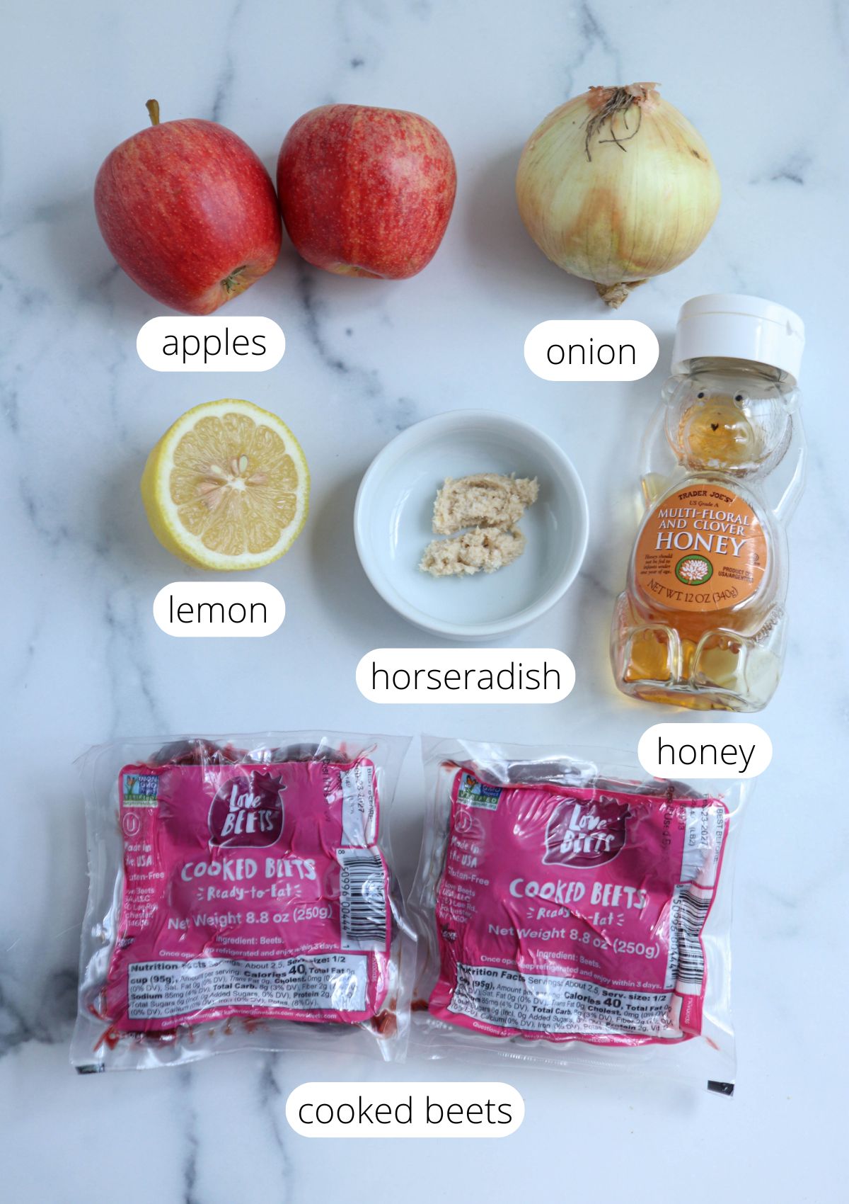 Top-down view of apples, an onion, a lemon half, horseradish, honey, and two packs of cooked beets on a white surface—all labeled—showcasing the fresh ingredients for a classic Polish beet salad.