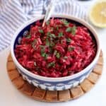 A bowl of vibrant Polish beet salad with shredded beets and apple, topped with chopped parsley. The bowl sits on a wooden trivet, with a striped cloth and a lemon half in the background. A fork is placed inside the bowl.