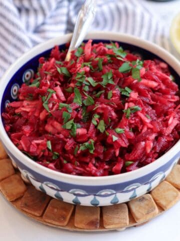 A bowl of vibrant Polish beet salad with shredded beets and apple, topped with chopped parsley. The bowl sits on a wooden trivet, with a striped cloth and a lemon half in the background. A fork is placed inside the bowl.