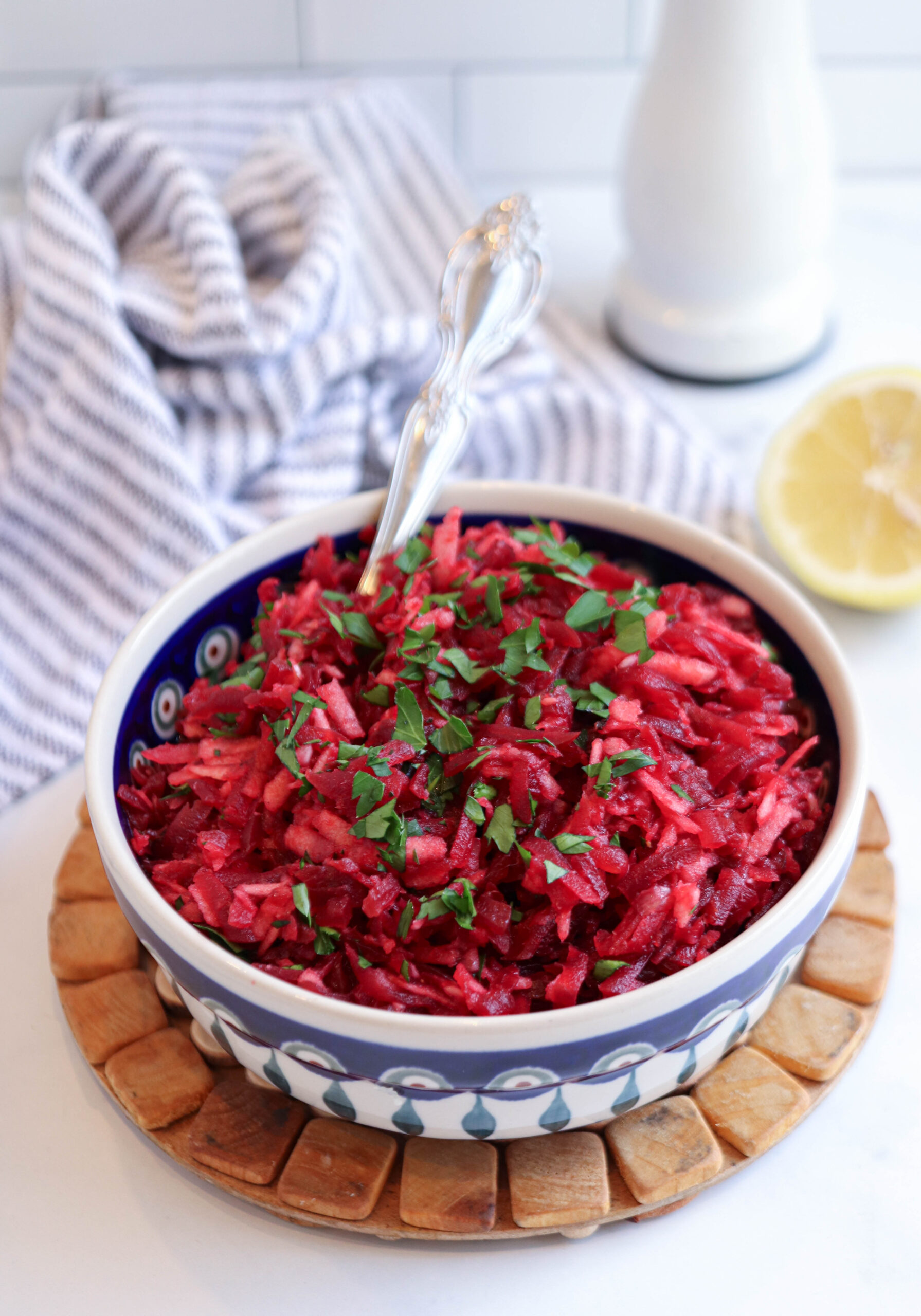 A bowl of vibrant Polish beet salad, garnished with chopped parsley and a fork nestled in the bowl, sits on a wooden trivet. A striped cloth and half a lemon are visible in the background.