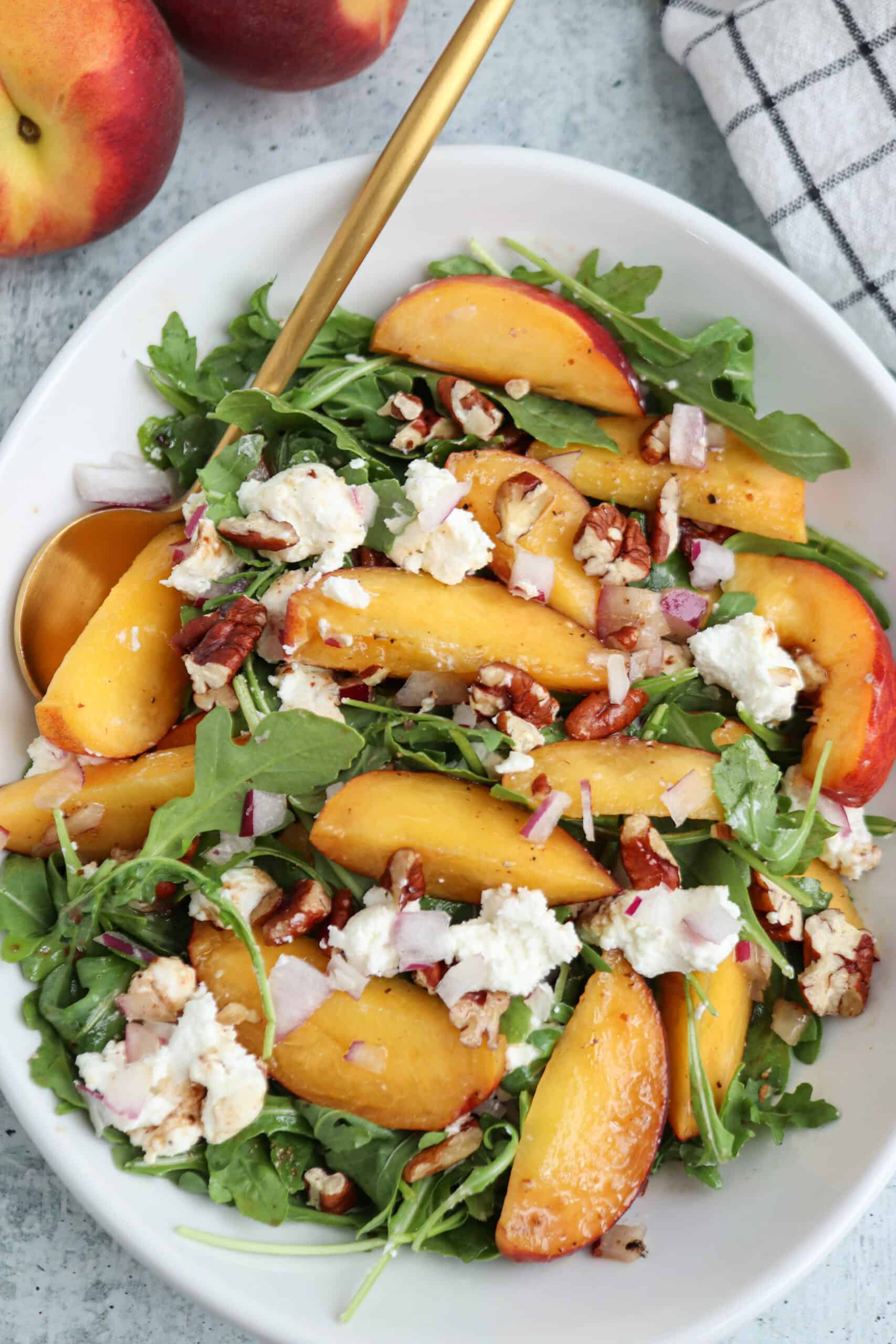 A fresh salad with arugula, sliced peaches, goat cheese, chopped pecans, and red onion is served in a white dish with a gold spoon. Two whole peaches and a checkered napkin are nearby, making it the perfect vibrant side to accompany hearty chicken parm meatballs for a balanced meal.