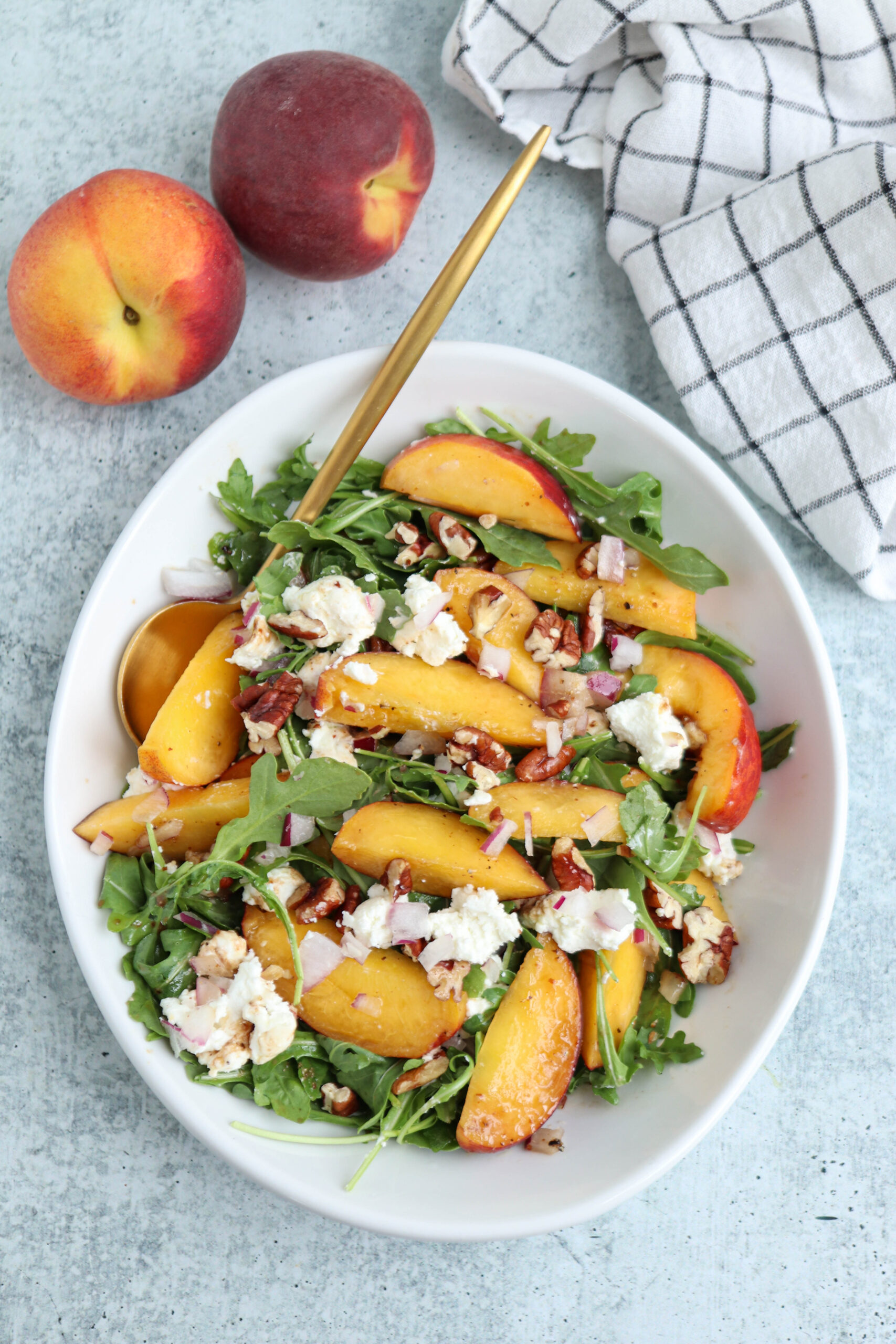 A bowl of arugula salad with sliced peaches, crumbled cheese, chopped nuts, and a gold fork sits beside two whole peaches and a checkered cloth on a light surface, making a vibrant pairing for hearty chicken parm meatballs served alongside.