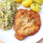 A plate with breaded fried cutlet, boiled potatoes sprinkled with dill, and sautéed cabbage with bacon pieces.