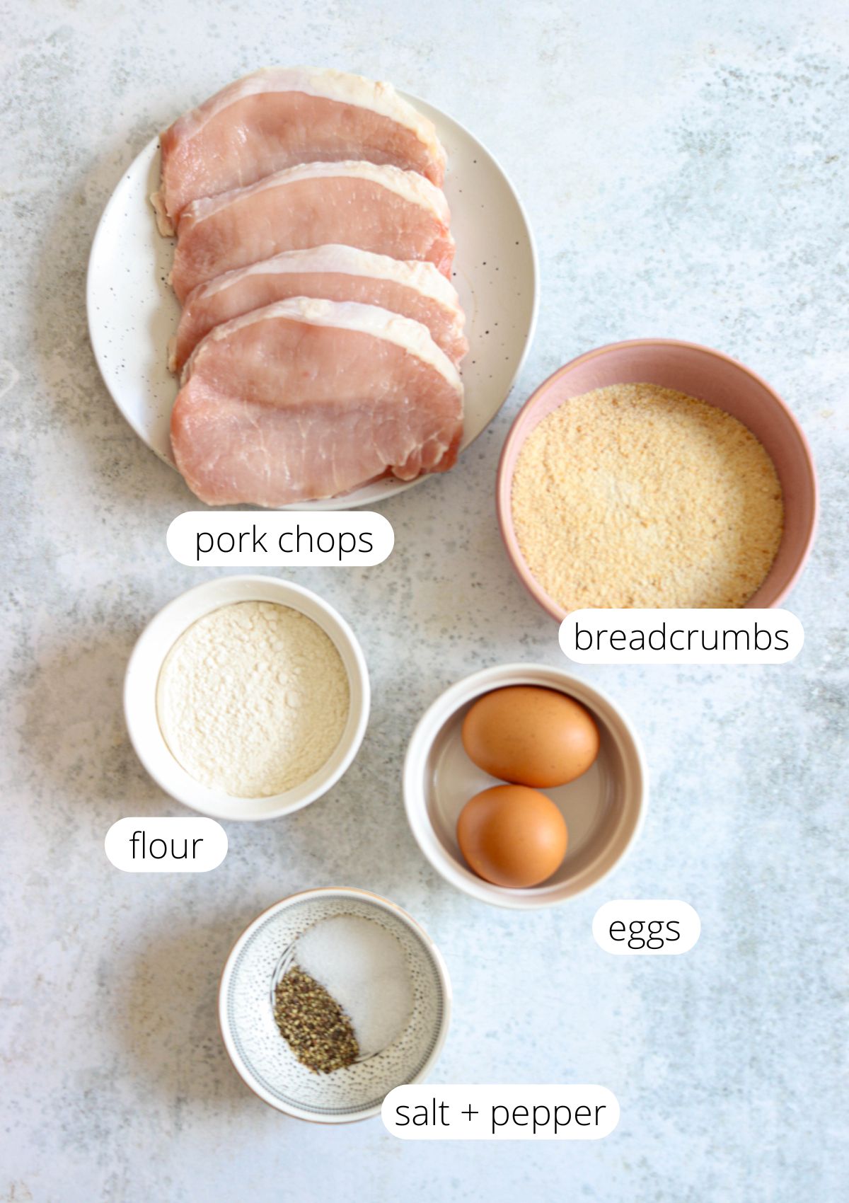 Five raw pork chops on a plate, bowls of breadcrumbs, flour, two eggs, and a small bowl with salt and pepper—all labeled on a light background.