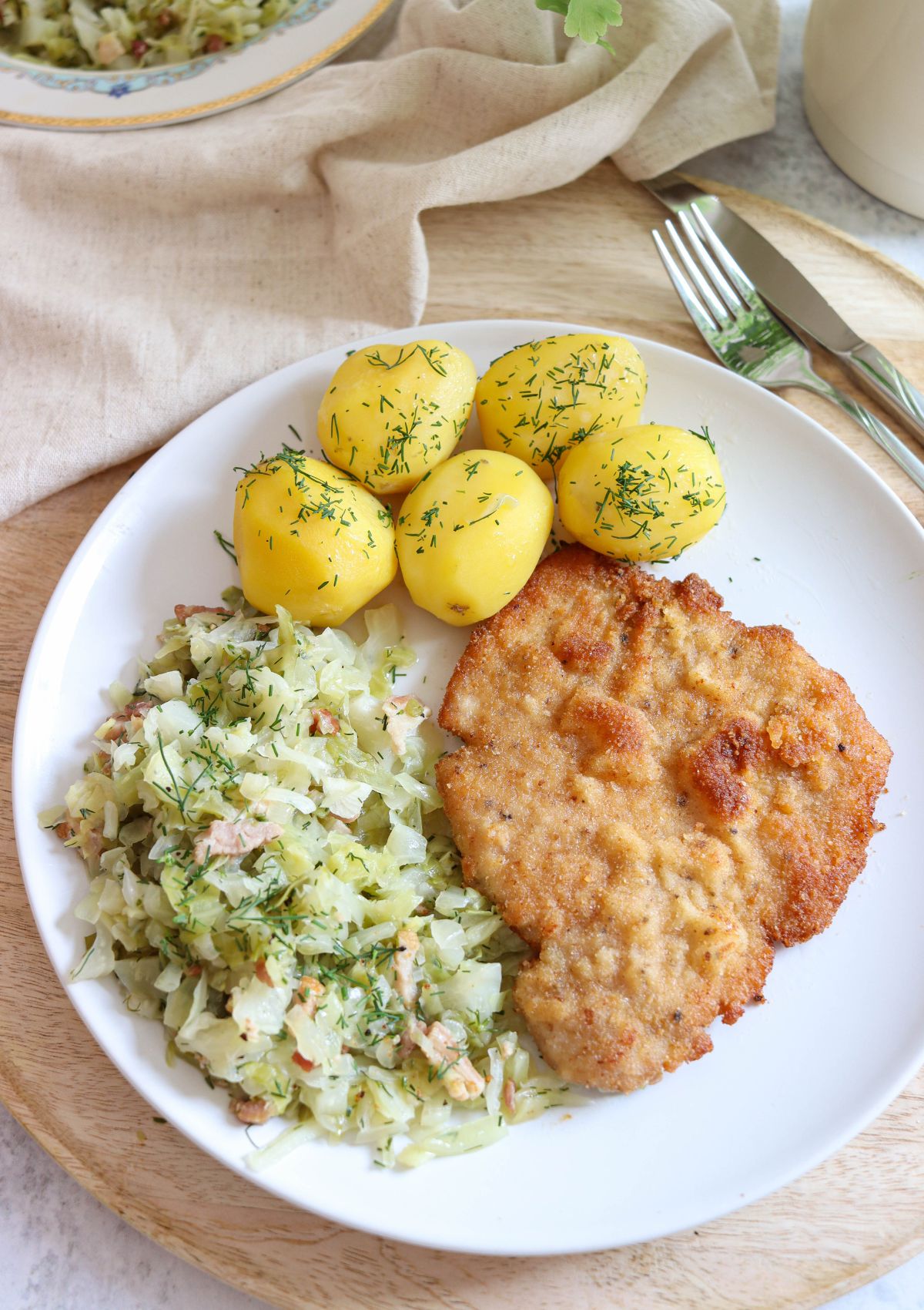 A white plate with kotlet schabowy, boiled potatoes topped with dill, and cooked cabbage mixed with bacon, set on a wooden tray with a fork and knife nearby.