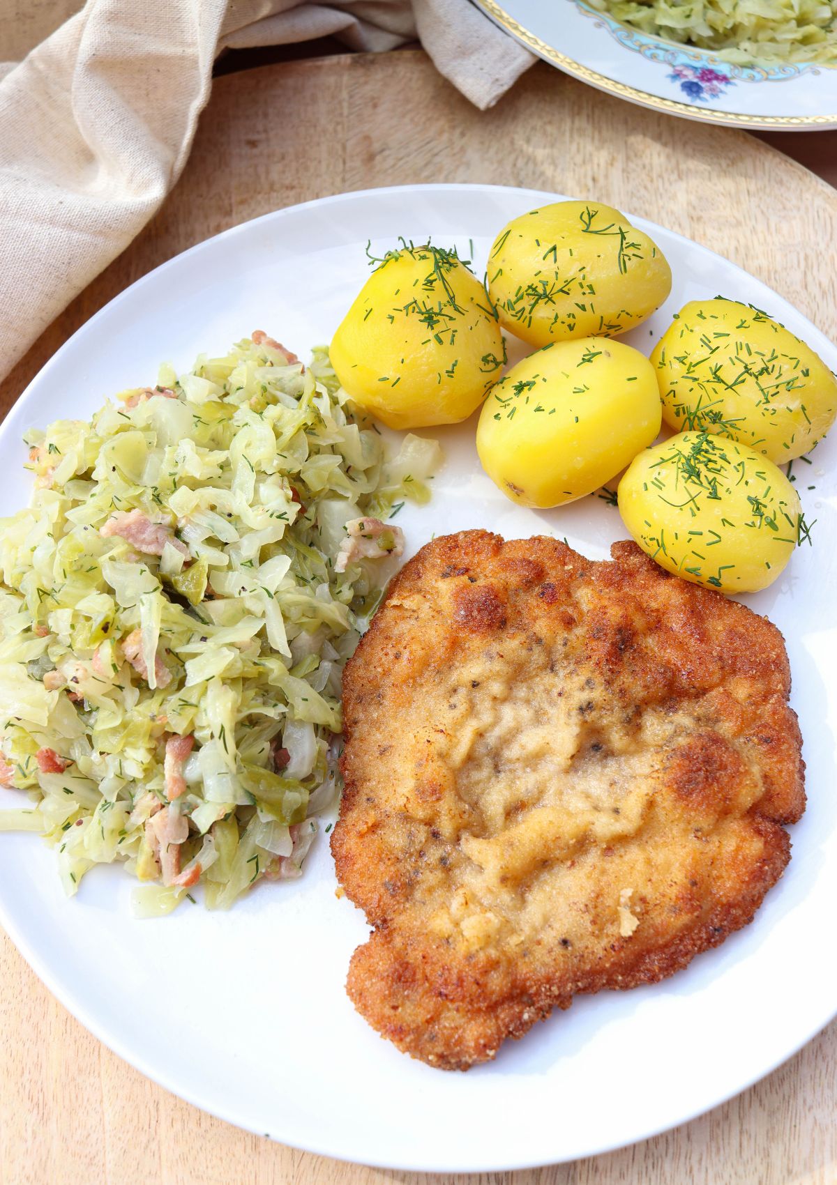 A plate with breaded kotlet schabowy, boiled potatoes topped with dill, and a side of cabbage mixed with bacon bits, all served on a white dish.