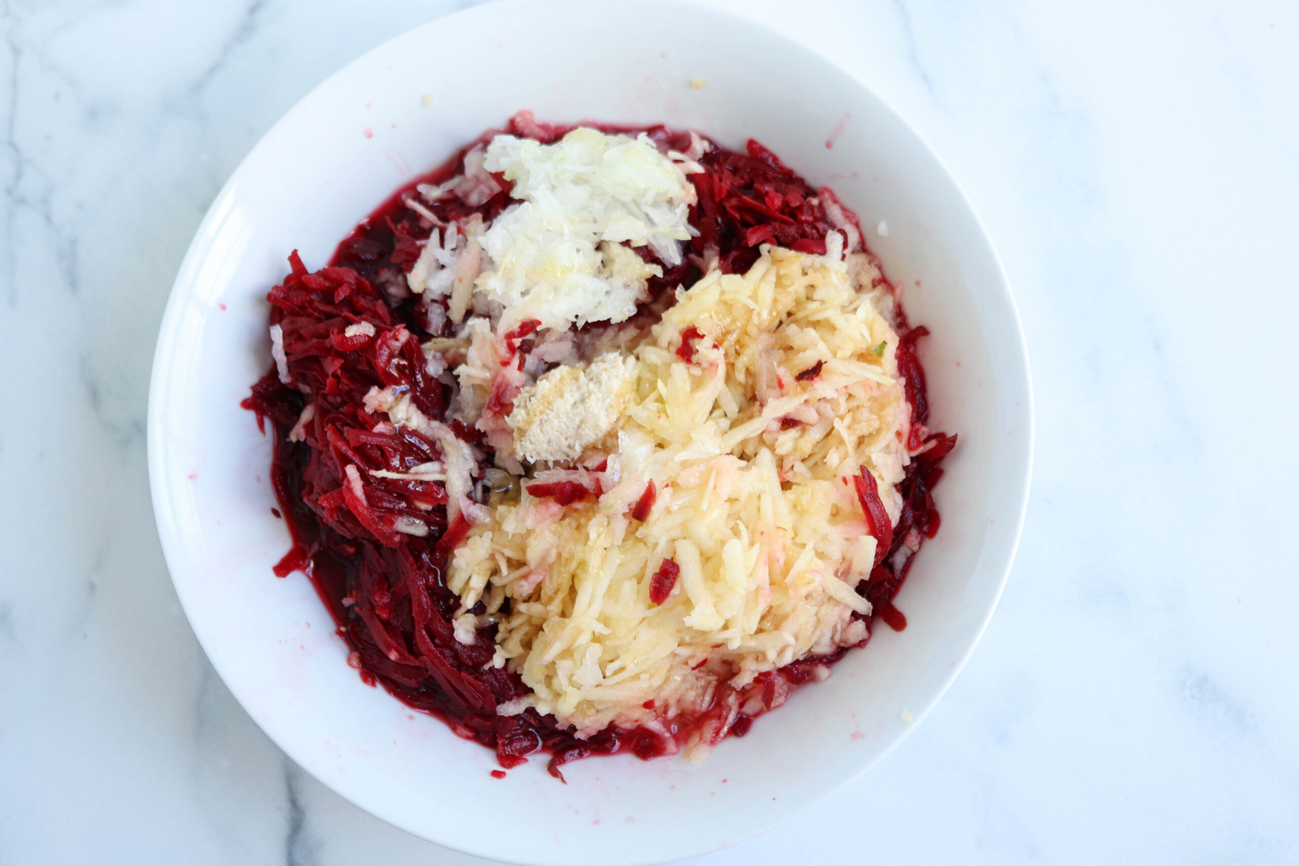 A white bowl containing Polish beet salad made with grated raw beets, potatoes, and onions on a white marble surface.
