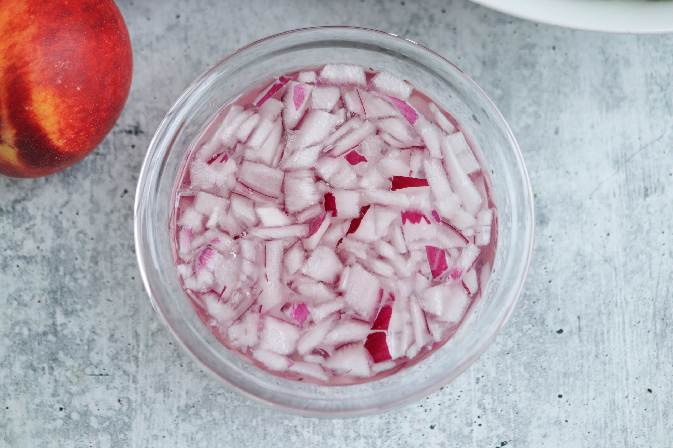 A glass bowl filled with finely chopped red onions soaking in liquid, placed on a light gray surface. A red and yellow fruit is partially visible to the side, adding a pop of color—perfect ingredients for topping fresh chicken parm meatballs.