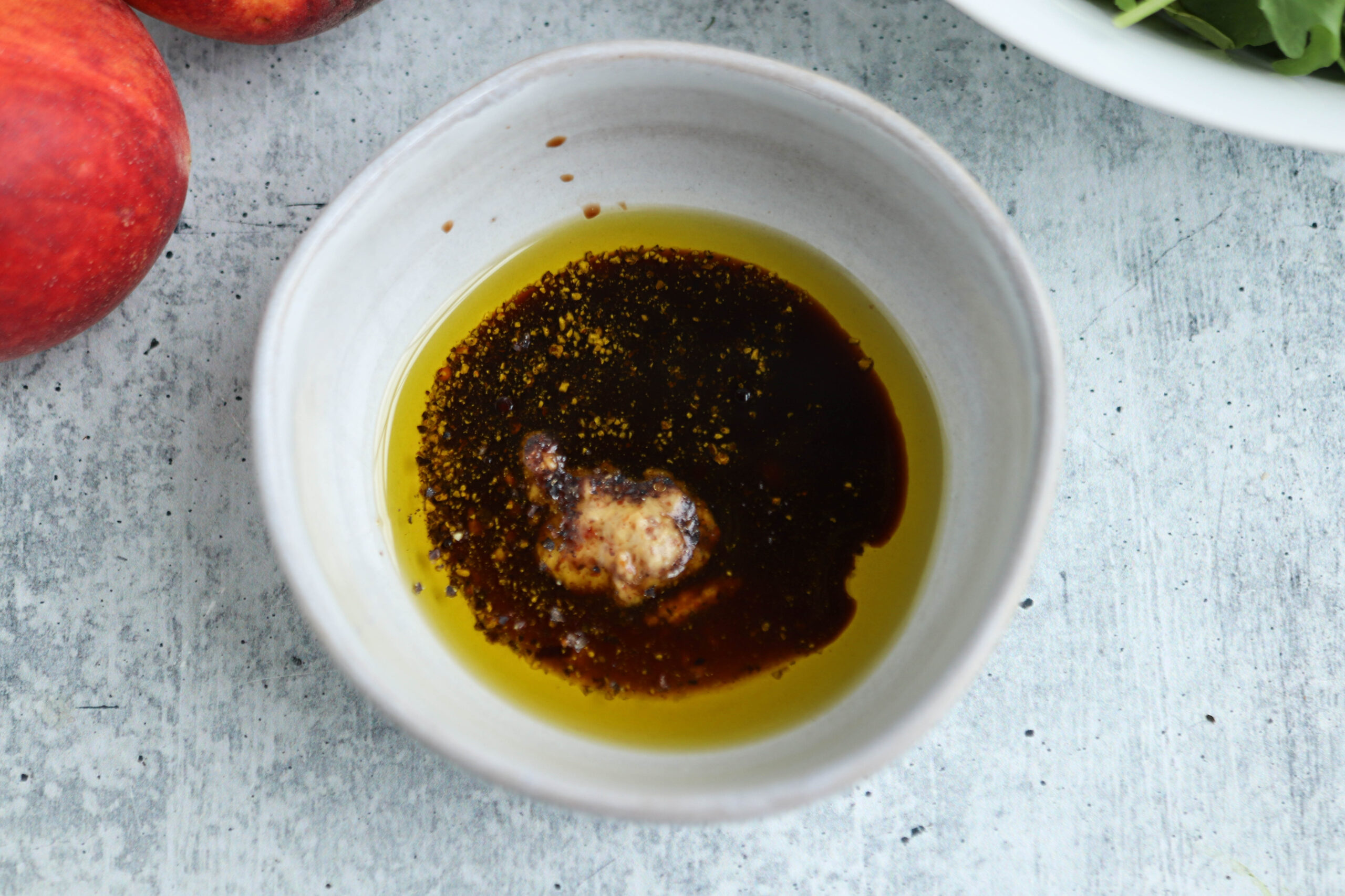 A small ceramic bowl contains olive oil, balsamic vinegar, and a dollop of mustard, likely being prepared for a salad dressing to accompany chicken parm meatballs. The background is a gray textured surface with part of a peach and leafy greens visible.