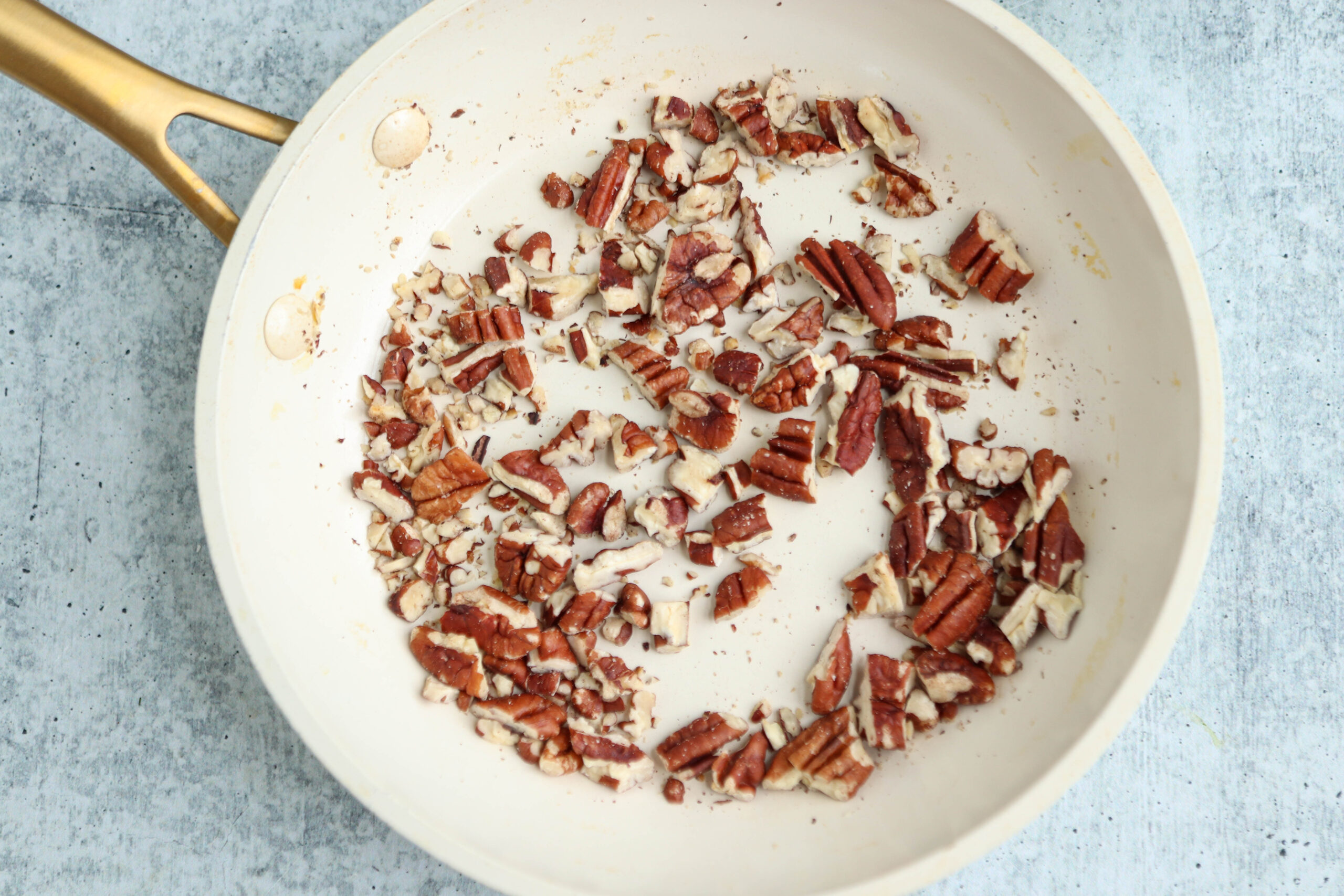 Chopped pecans being toasted in a white frying pan with a gold handle, placed on a light gray textured surface, ready to add a delicious crunch to your chicken parm meatballs.