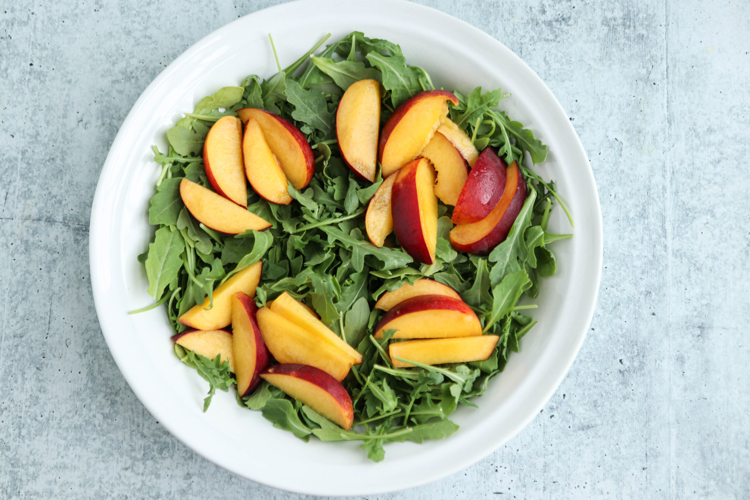 A white bowl filled with fresh arugula leaves and savory chicken parm meatballs, topped with sliced peaches, placed on a light gray textured surface.