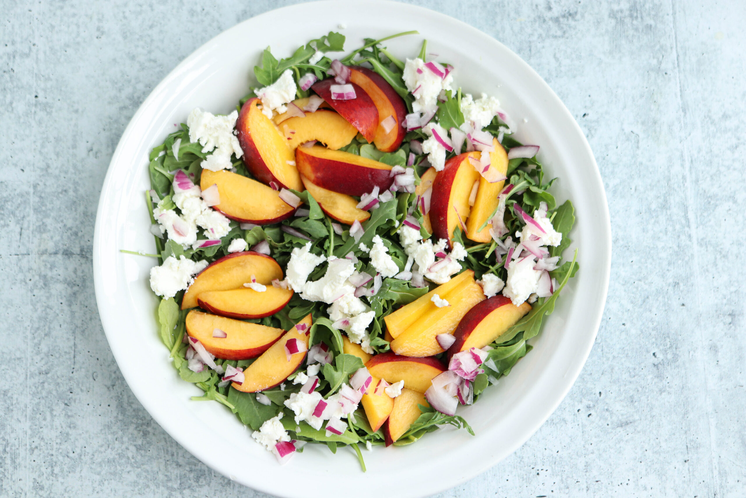 A white plate with a salad of arugula, sliced peaches, crumbled feta cheese, and chopped red onions is paired perfectly with savory chicken parm meatballs on a light gray surface.