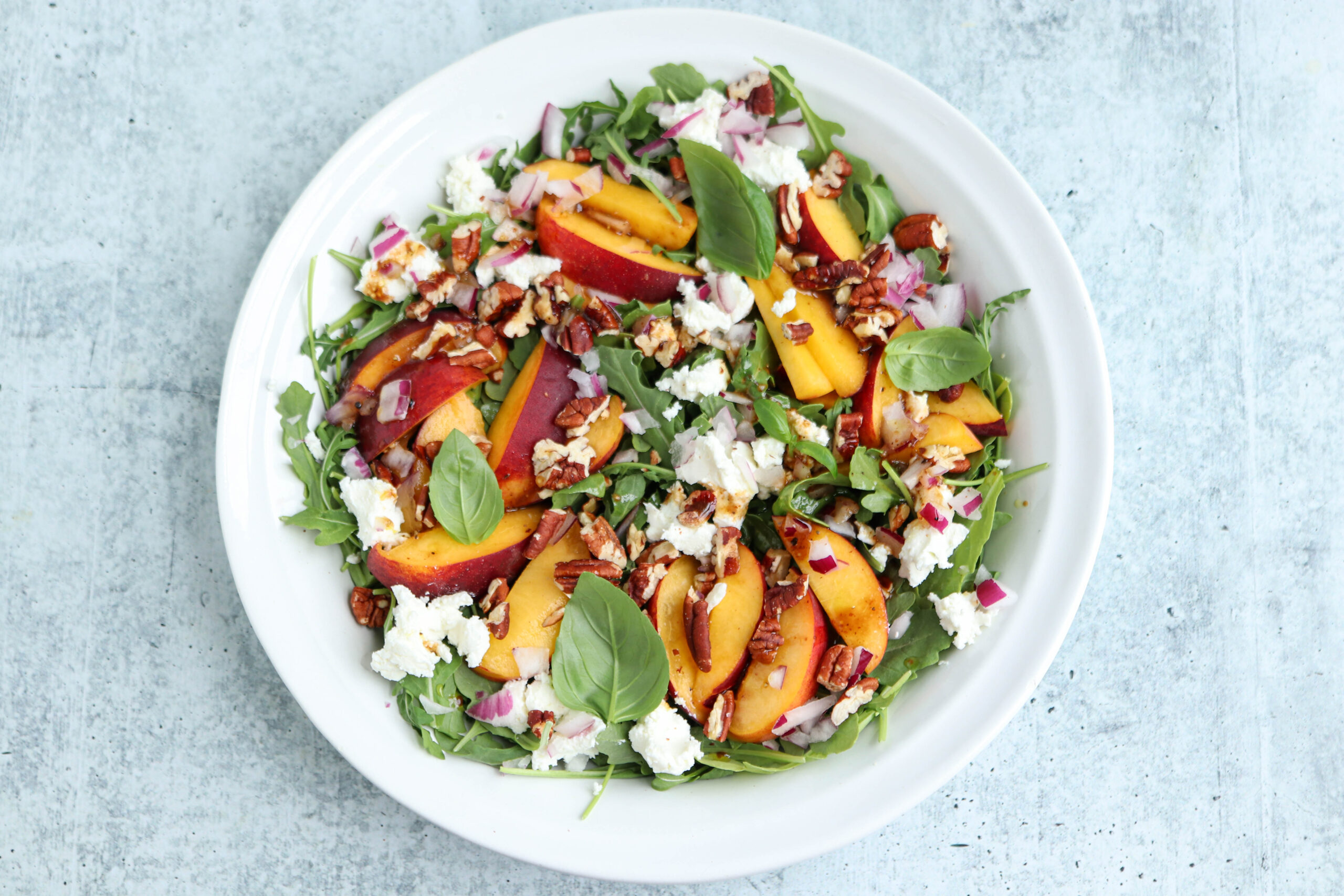 A white bowl filled with arugula, sliced peaches, crumbled feta, chopped red onions, pecans, chicken parm meatballs, and fresh basil leaves on a light gray background.