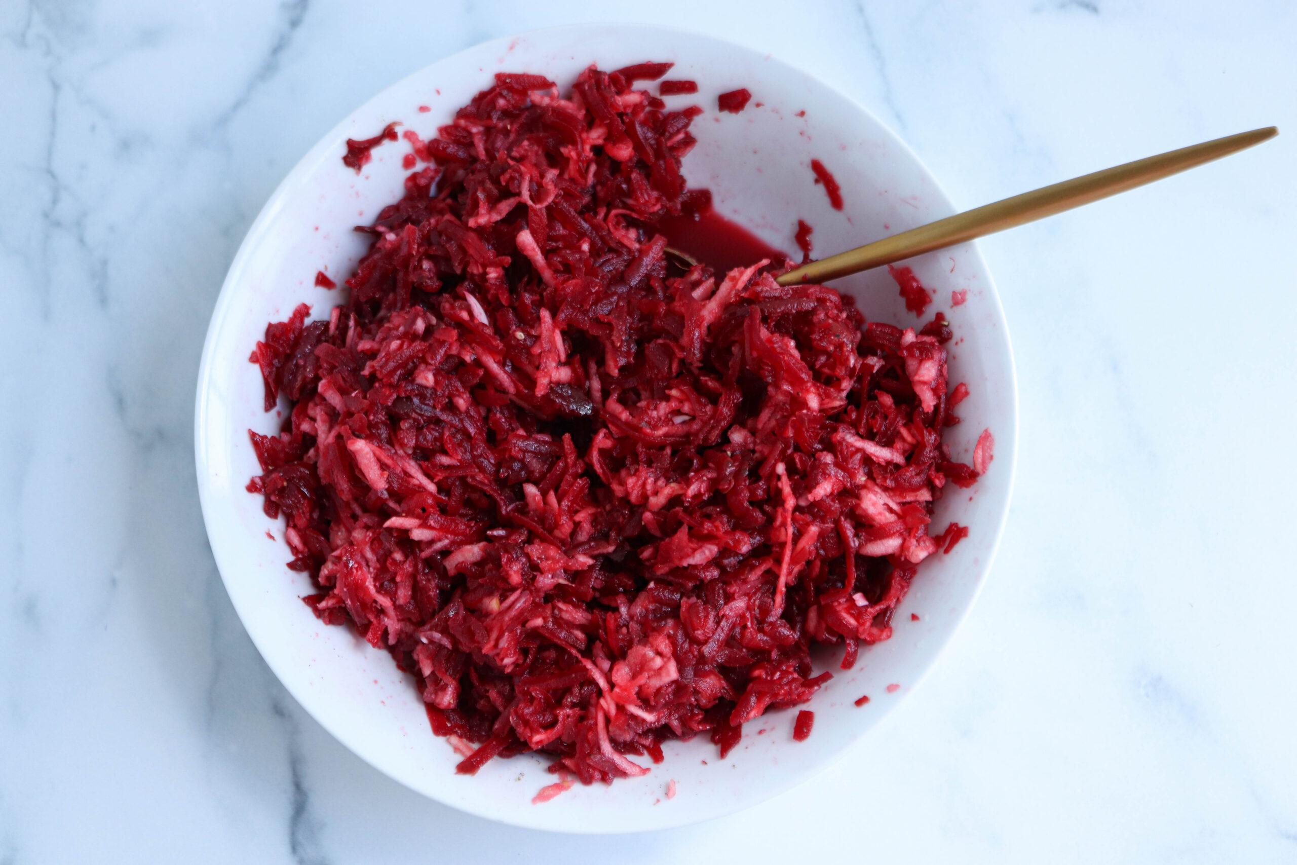 A white bowl filled with shredded red and white vegetables, likely beetroot and cabbage, sits atop a white marble surface—a classic Polish beet salad—with a golden spoon resting inside.