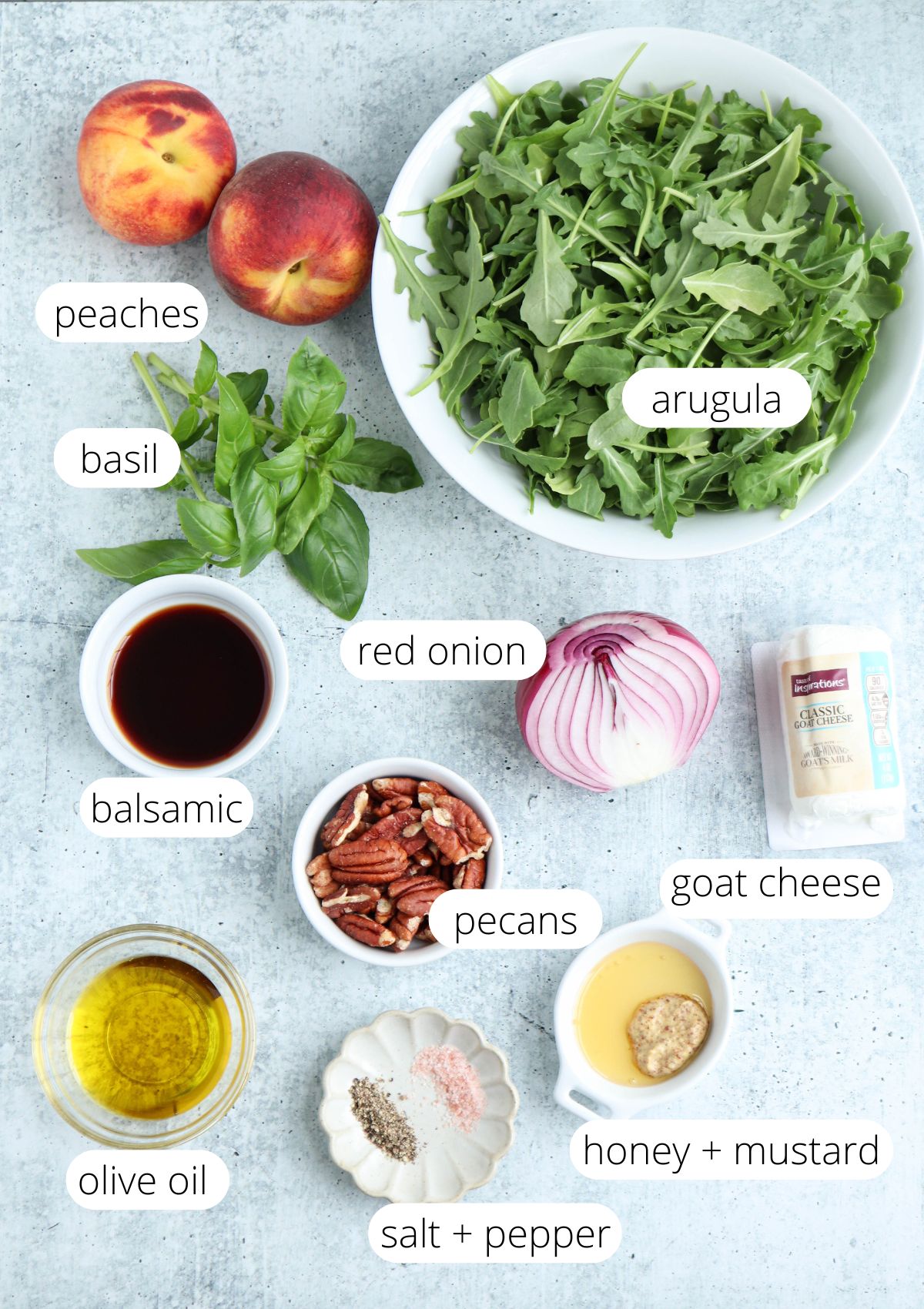 Overhead view of salad ingredients: arugula, peaches, basil, red onion, goat cheese, pecans, balsamic vinegar, olive oil, honey, mustard, and salt and pepper—each labeled with text—for a vibrant summer dish that pairs perfectly with chicken parm meatballs.