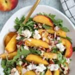 A bowl of peach arugula salad is topped with sliced peaches, creamy goat cheese, and chopped nuts. A gold fork rests in the bowl. Two whole peaches and a striped napkin are nearby. Text reads: Fresh Peach Arugula Salad with Goat Cheese and Nuts.