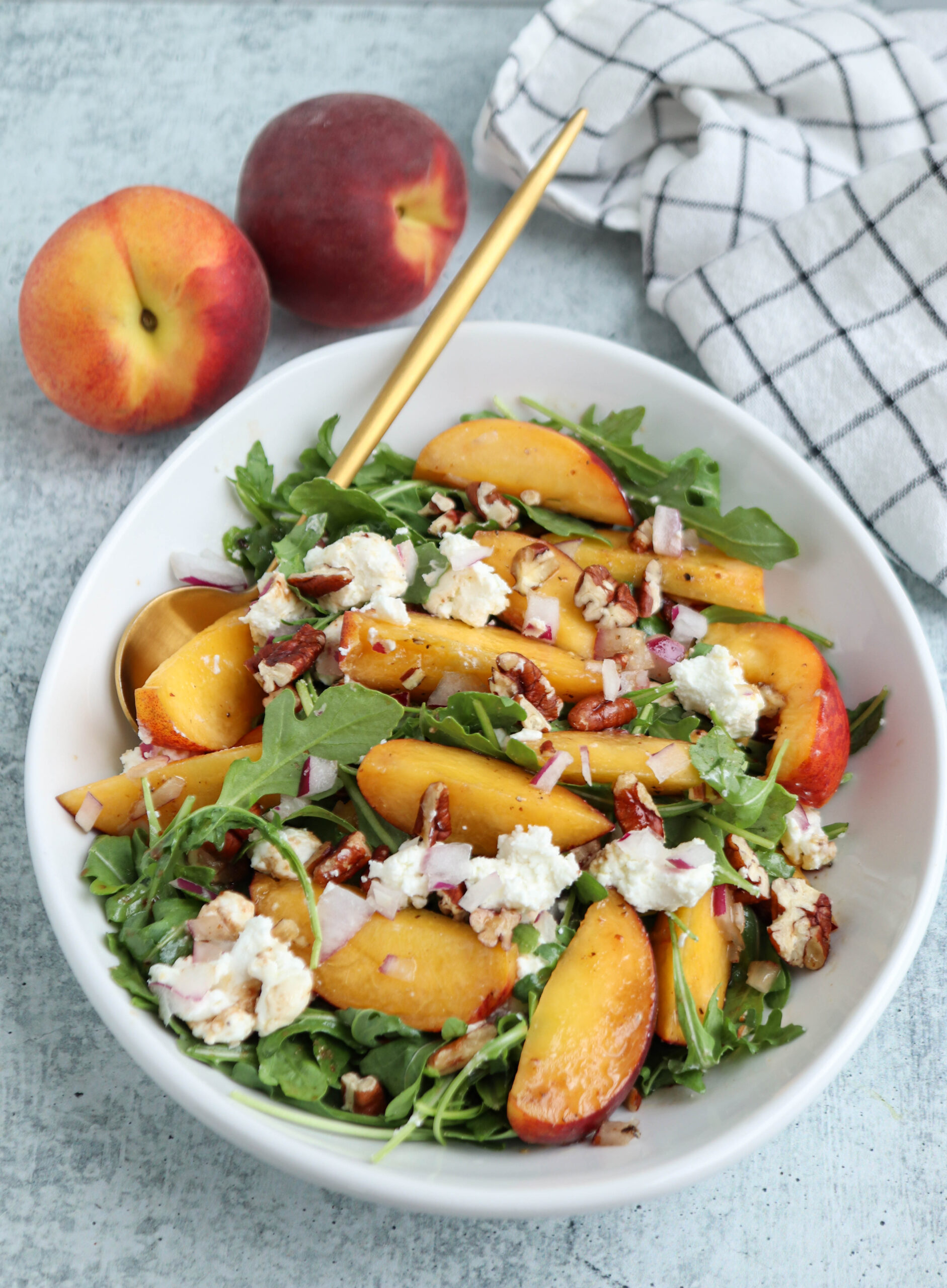 A bowl of arugula salad topped with sliced peaches, crumbled cheese, pecans, and red onion sits invitingly on the table. A gold fork rests in the bowl, ready to enjoy alongside hearty chicken parm meatballs for a balanced meal. Two whole peaches and a checkered napkin are nearby.