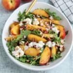 A white bowl of arugula salad topped with sliced peaches, crumbled cheese, chopped pecans, and diced red onions sits next to a plate of savory chicken parm meatballs, with a gold spoon on the side and a checked napkin nearby.