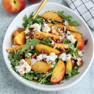A white bowl of arugula salad topped with sliced peaches, crumbled cheese, chopped pecans, and diced red onions sits next to a plate of savory chicken parm meatballs, with a gold spoon on the side and a checked napkin nearby.