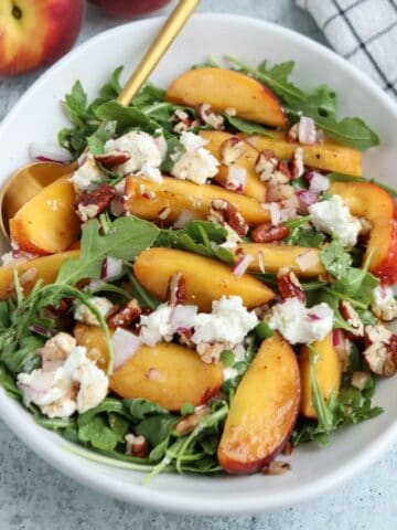 A white bowl of arugula salad topped with sliced peaches, crumbled cheese, chopped pecans, and diced red onions sits next to a plate of savory chicken parm meatballs, with a gold spoon on the side and a checked napkin nearby.
