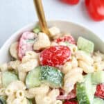 A bowl of creamy tomato cucumber pasta salad with cucumber slices, cherry tomatoes, and a fork, on a light surface. The text above reads, Creamy Tomato Cucumber Pasta Salad—Lighter Macaroni Salad.