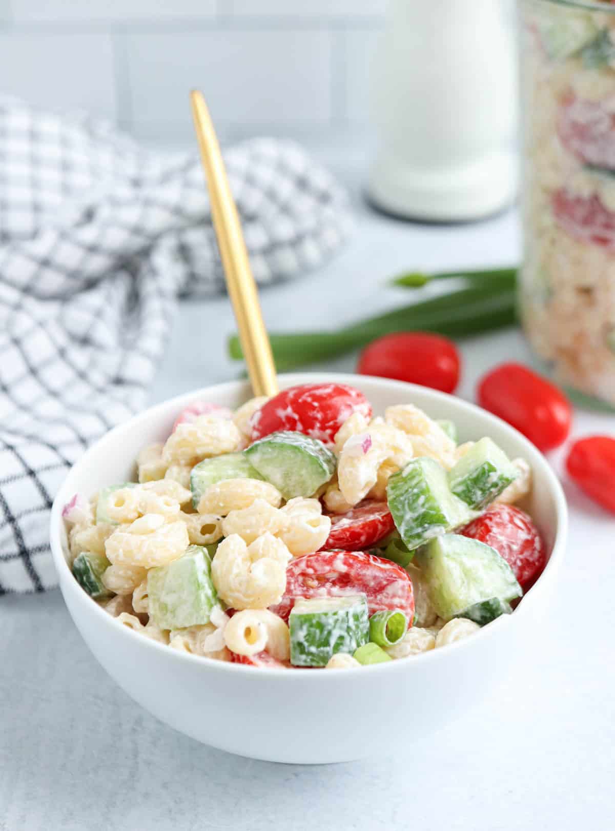 A white bowl of creamy tomato cucumber pasta salad with sliced cucumbers, grape tomatoes, and green onions sits on a light surface. A gold fork is in the bowl. A checkered cloth and tomatoes are in the background.