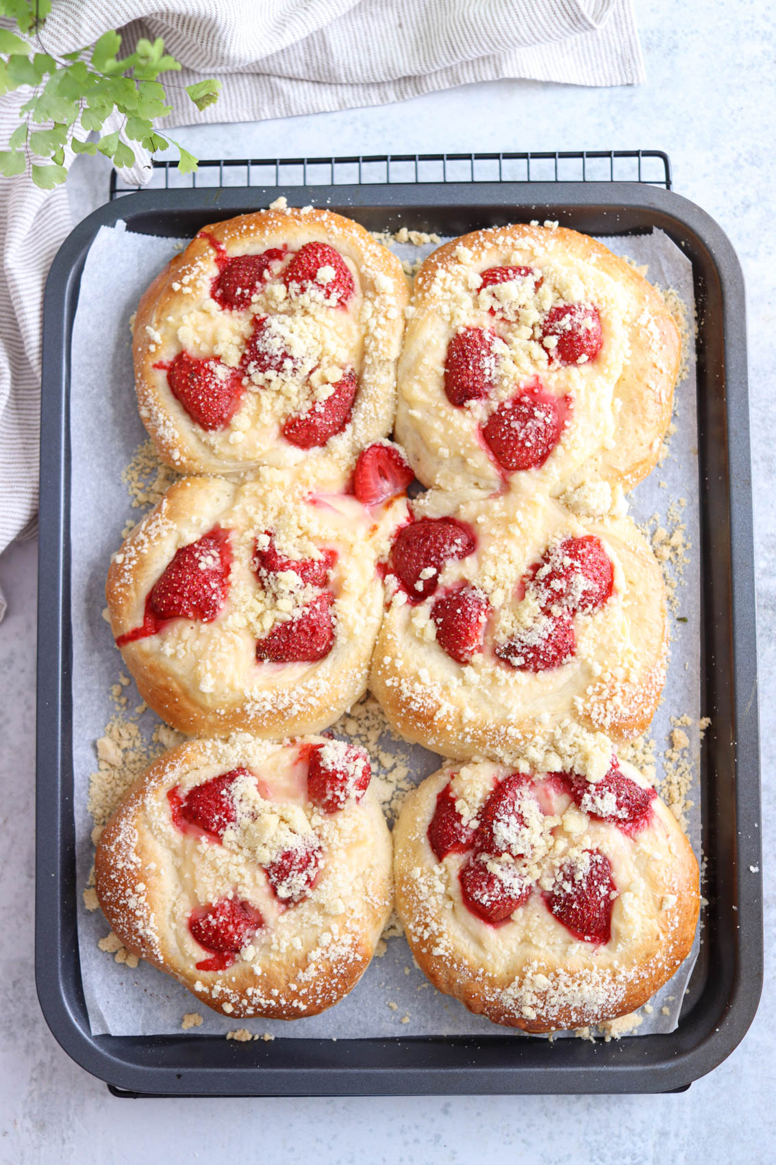 Six round Polish sweet rolls, or drożdżówki, topped with fresh strawberries and crumbly streusel are arranged in a baking tray lined with parchment paper. A beige cloth and a small green plant are partially visible in the background.
