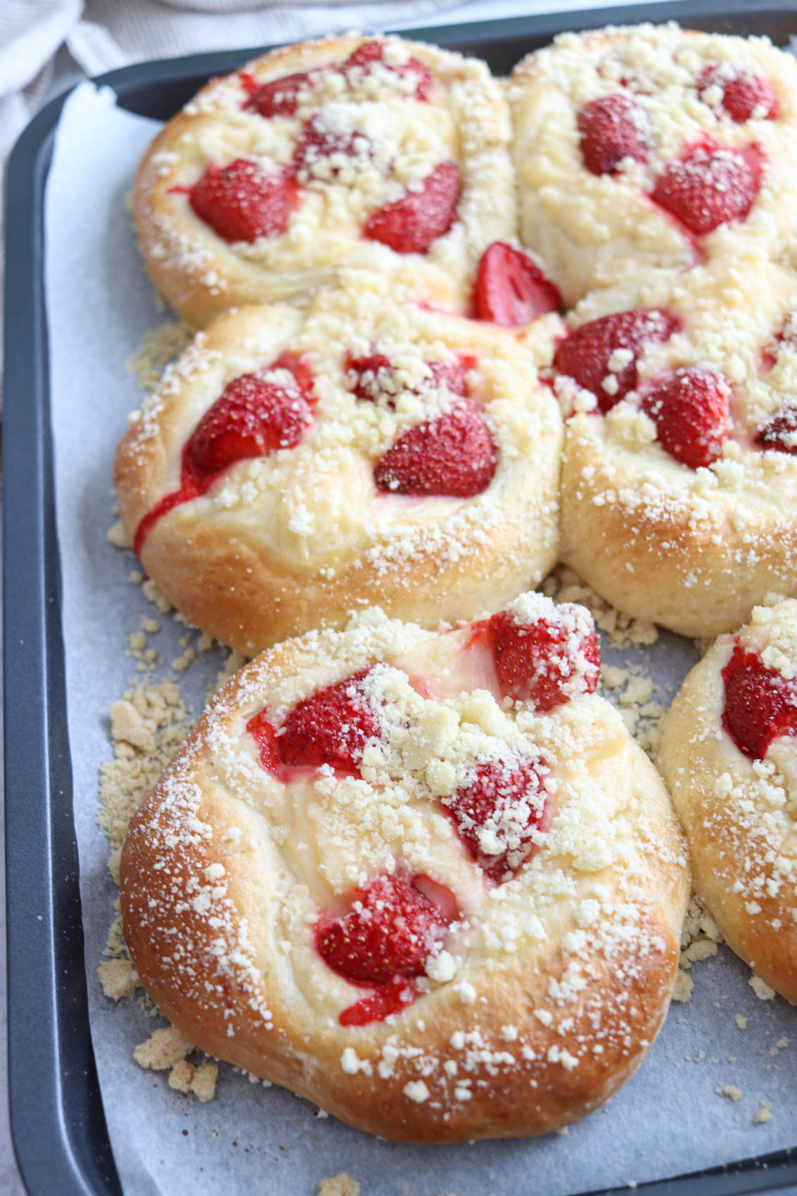 Six round Polish sweet rolls drożdżówki topped with sliced strawberries and crumbly streusel sit on a parchment-lined baking tray. The pastries are golden brown with a creamy filling visible under the fruit and crumbs.