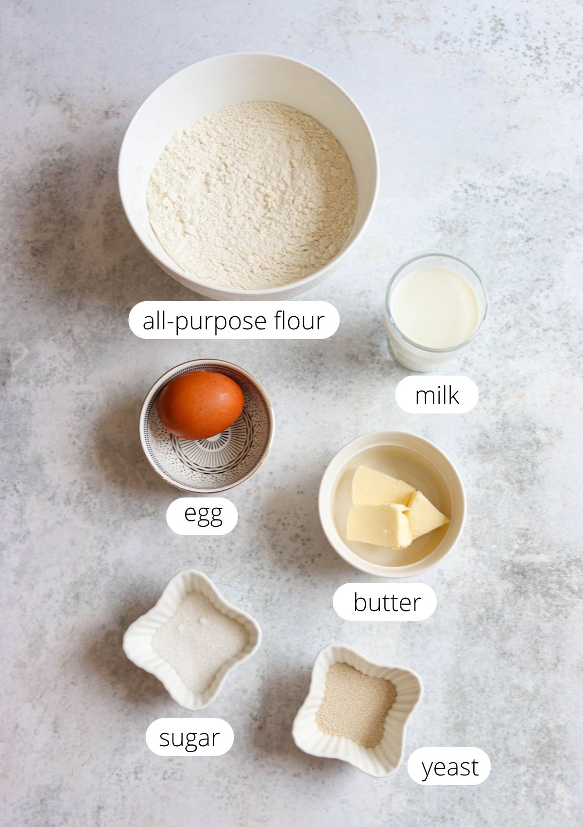 A flat lay of labeled baking ingredients for Polish sweet rolls drożdżówki: a bowl of all-purpose flour, a glass of milk, an egg, butter, sugar, and yeast, all arranged on a light surface.