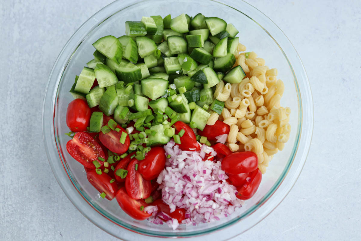 A glass bowl with chopped cucumbers, halved grape tomatoes, diced red onion, cooked pasta, and sliced green onions—perfectly arranged in separate sections on a light surface—makes for a fresh and colorful tomato cucumber pasta salad.