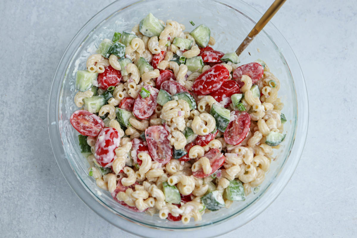 A glass bowl filled with a delicious tomato cucumber pasta salad, featuring macaroni pasta, cherry tomatoes, crisp cucumbers, green onions, and a creamy dressing, with a spoon inside.