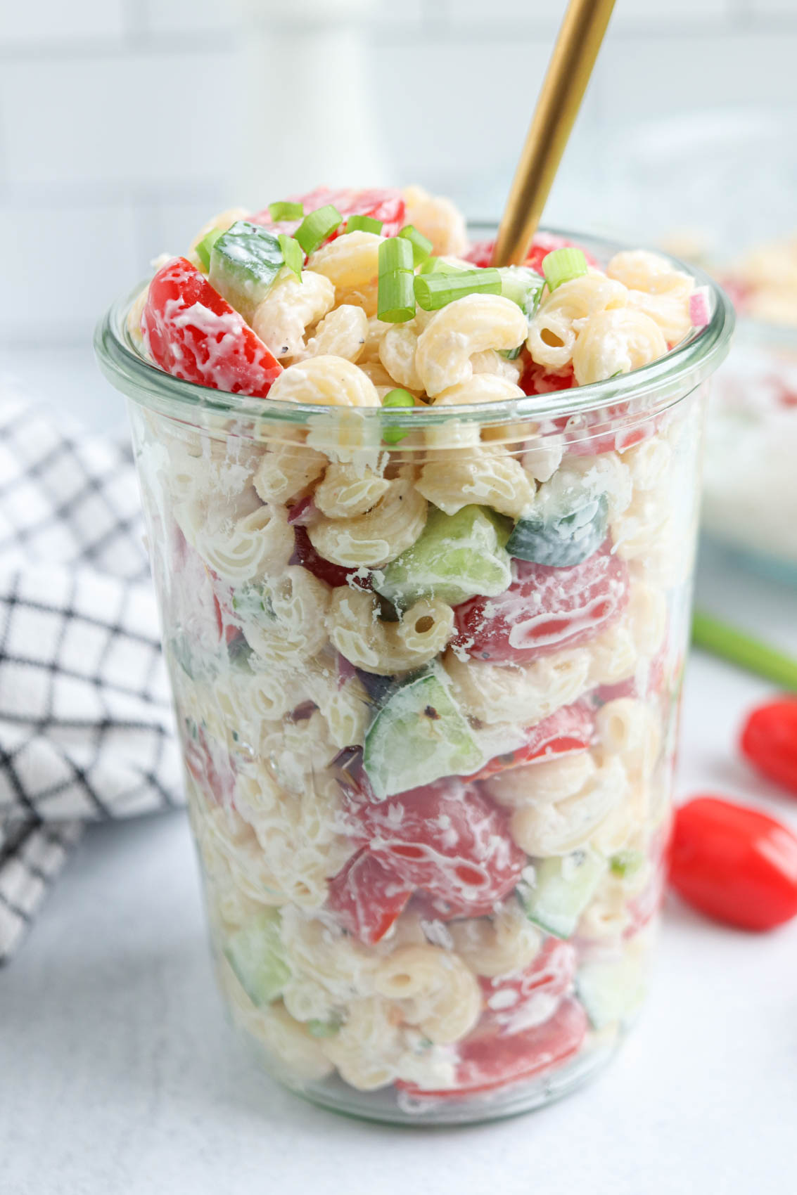 A glass jar filled with creamy tomato cucumber pasta salad, featuring elbow macaroni, chopped cucumbers, tomatoes, and green onions, with a gold fork inside. A black-and-white towel and cherry tomatoes are in the background.