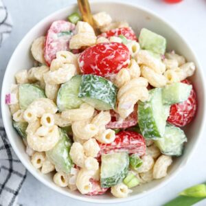 A bowl of creamy tomato cucumber pasta salad with elbow macaroni, sliced cucumbers, cherry tomatoes, chopped green onions, and red onions, all mixed with a white dressing.