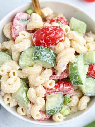 A bowl of creamy tomato cucumber pasta salad with elbow macaroni, sliced cucumbers, cherry tomatoes, chopped green onions, and red onions, all mixed with a white dressing.