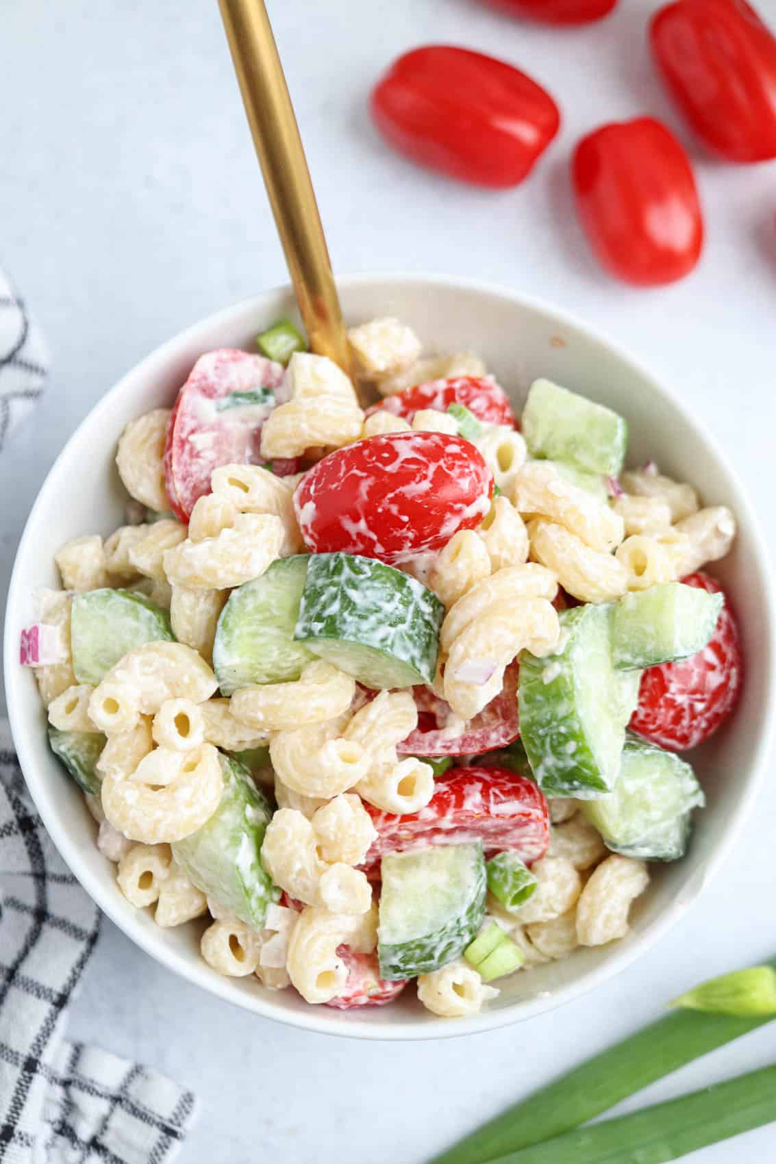 A bowl of creamy tomato cucumber pasta salad with macaroni, cherry tomatoes, cucumber, and a gold fork. Some cherry tomatoes and green onions are on the side.