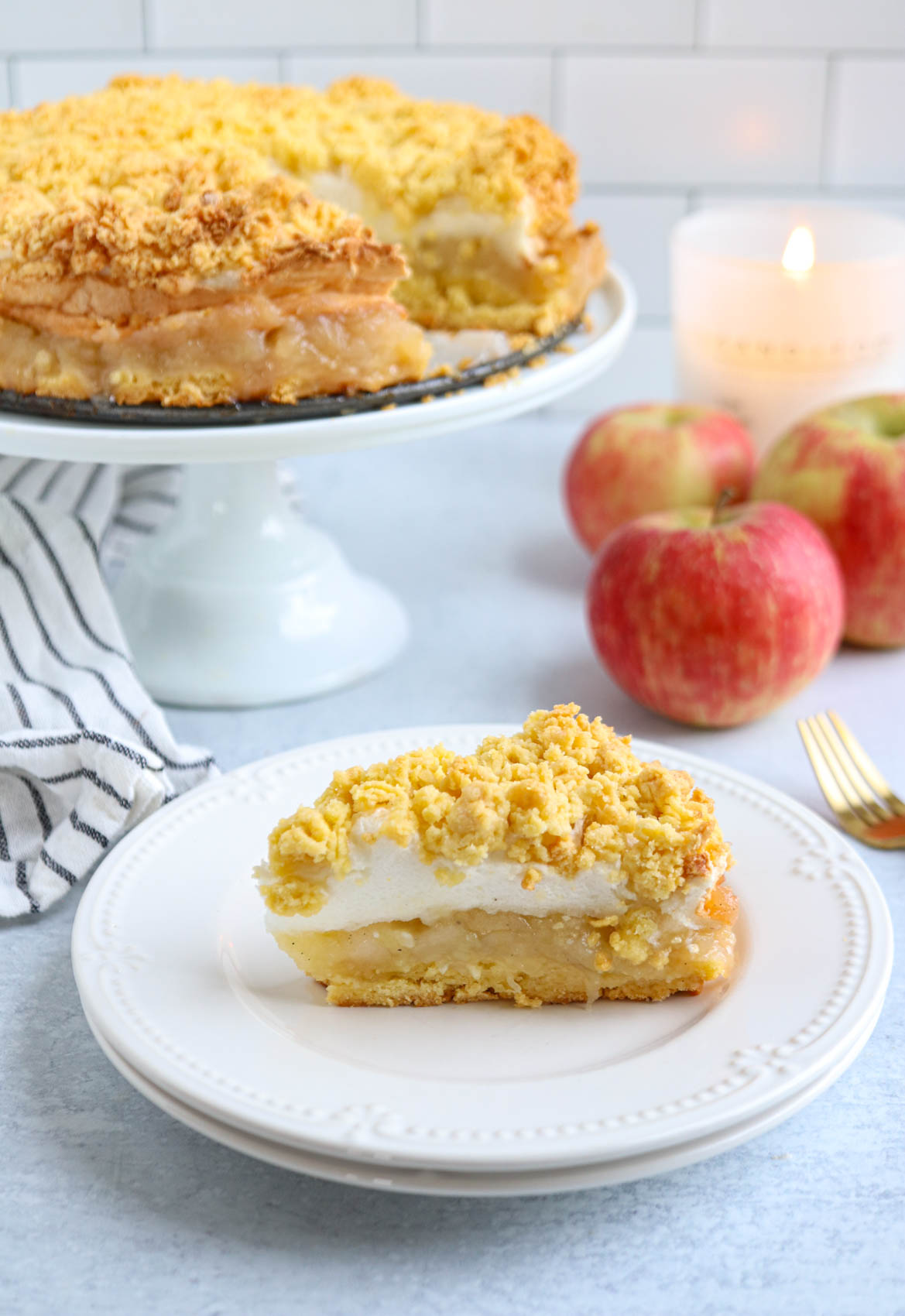 A slice of layered Polish apple pie with crumb topping sits on a white plate; the rest of the dessert is on a cake stand in the background, with red apples and a lit candle nearby.