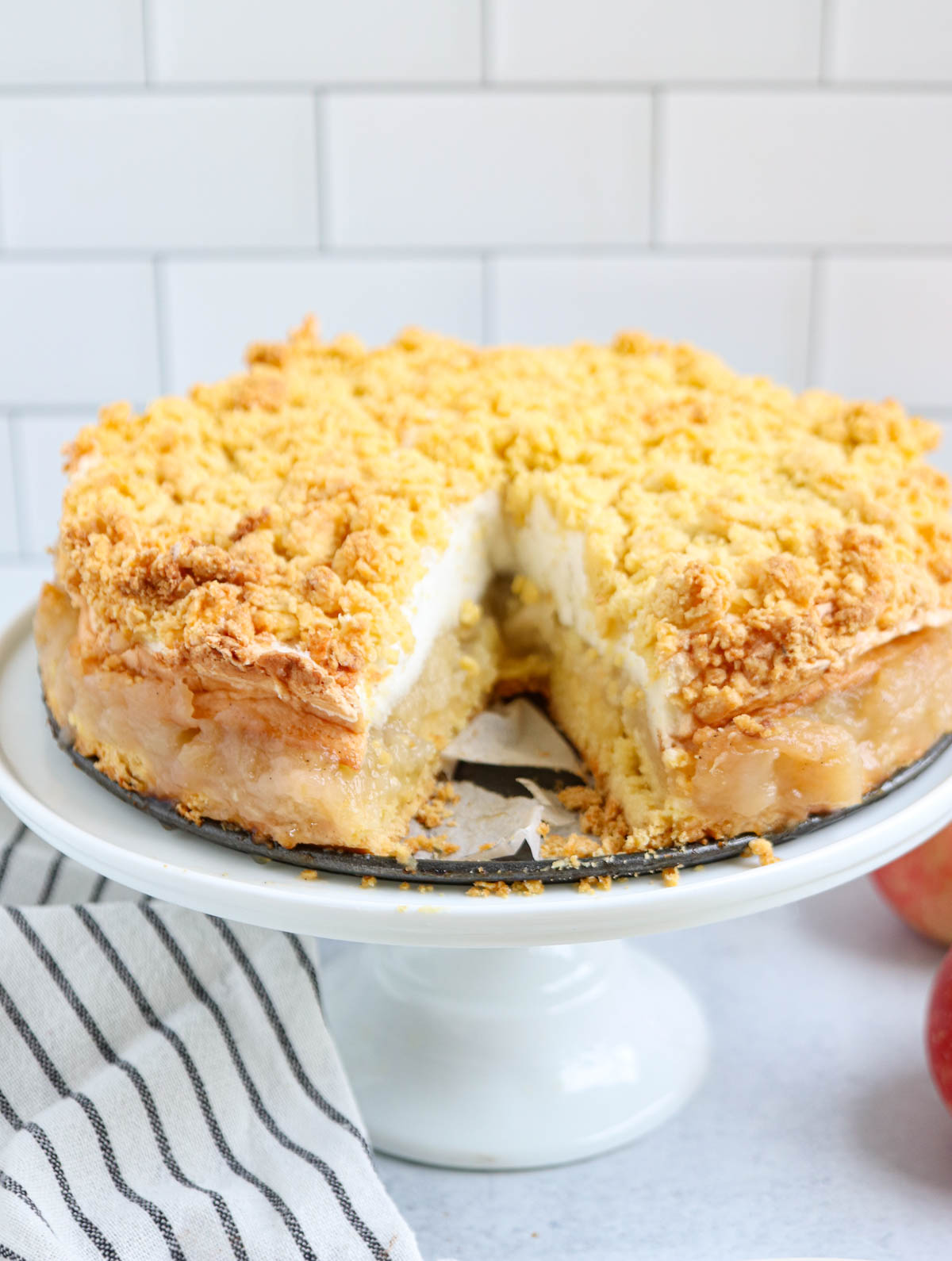 A crumb-topped Polish apple pie with a slice missing, displayed on a white cake stand. A striped towel is next to the stand, and the background features a white tiled wall.
