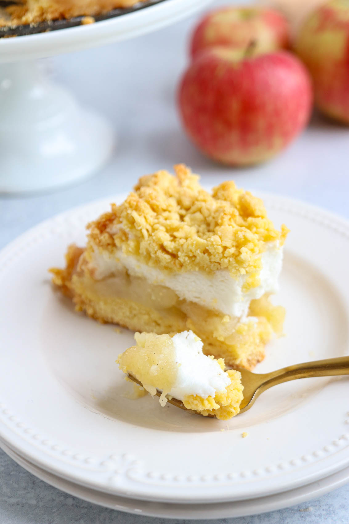 A slice of crumb-topped Polish apple pie with a layer of meringue sits on a white plate, next to a forkful of the dessert, with apples in the background.