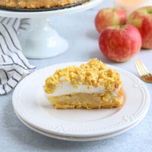 A slice of apple crumble pie with a crumb topping and meringue layer sits on a white plate; whole apples, a pie on a stand, and a bowl of sardine salad are in the background.