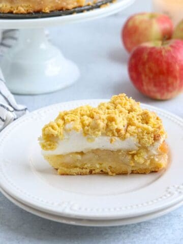 A slice of apple crumble pie with a crumb topping and meringue layer sits on a white plate; whole apples, a pie on a stand, and a bowl of sardine salad are in the background.