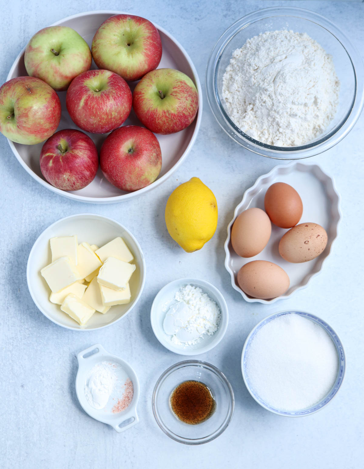 Top-down view of baking ingredients for a Polish apple pie: apples, flour, eggs, butter, lemon, sugar, vanilla, baking powder, cornstarch, and salt, all arranged in separate bowls and plates on a light surface.