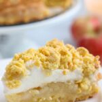 A slice of Polish apple pie Szarlotka with crumbly topping, soft apple filling, and a light layer is served on a white plate. In the background, a whole Polish apple pie and fresh apples are blurred for a cozy touch.